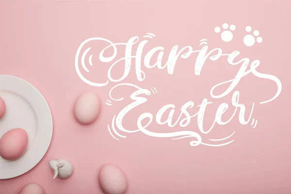 Top view of painted eggs and white decorative rabbit near plate on pink background with happy Easter lettering — Stock Photo