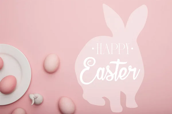 Top view of painted eggs and white decorative bunny near plate on pink background with happy Easter lettering — Stock Photo