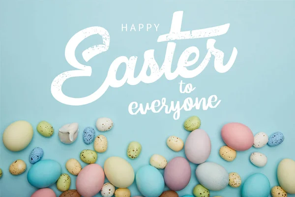 Top view of painted multicolored eggs scattered near decorative bunny on blue background with happy Easter to everyone lettering — Stock Photo