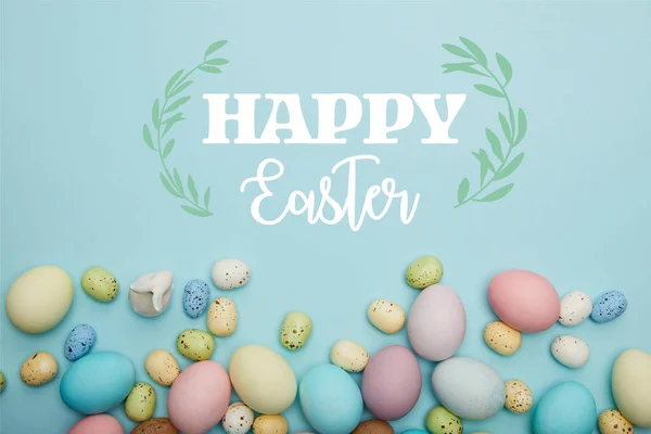 Top view of painted multicolored eggs scattered and decorative bunny on blue background with happy Easter lettering — Stock Photo