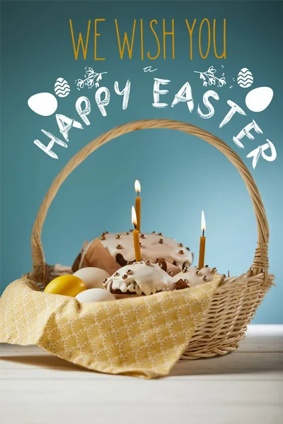 Traditional Easter cakes with burning candles and chicken eggs in wicker basket on blue background with we wish you happy easter lettering — Stock Photo