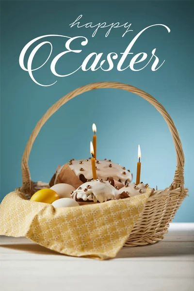 Traditional Easter cakes with burning candles and chicken eggs in wicker basket on blue background with happy easter lettering — Stock Photo
