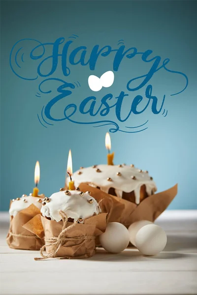 Traditional Easter cakes with burning candles and white chicken eggs on blue background with happy easter lettering — Stock Photo