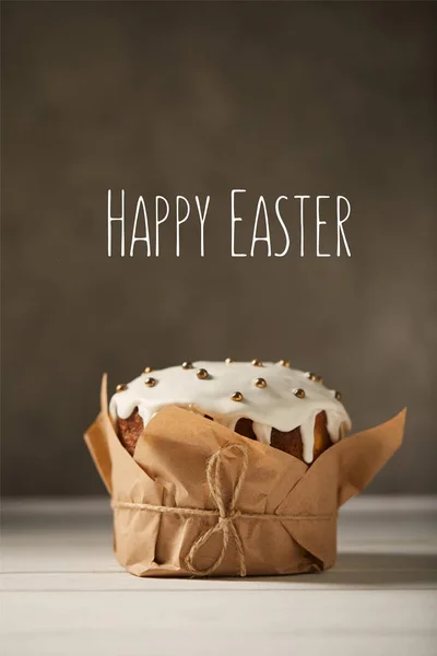Traditional decorated Easter cake in craft paper on white table with happy easter lettering on brown background — Stock Photo