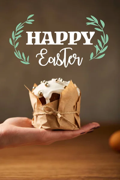 Cropped view of woman holding traditional Easter cake in craft paper with happy easter lettering above — Stock Photo