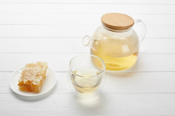 Tetera con té de hierbas orgánicas, vidrio y panal de abeja en mesa de madera blanca - foto de stock