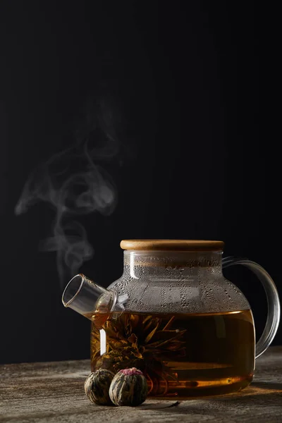 Tetera transparente con bolas de té y té en flor sobre mesa de madera aislada en negro con espacio de copia - foto de stock