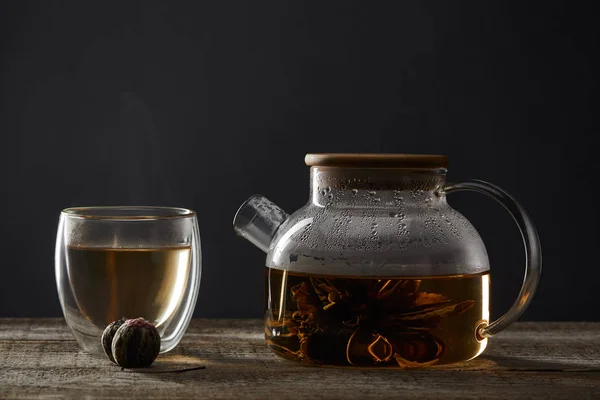 Tetera transparente y vidrio con té en flor sobre mesa de madera aislada en negro - foto de stock