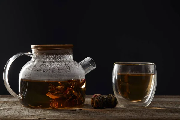 Transparent teapot and glass with blooming tea on wooden table isolated on black — Stock Photo