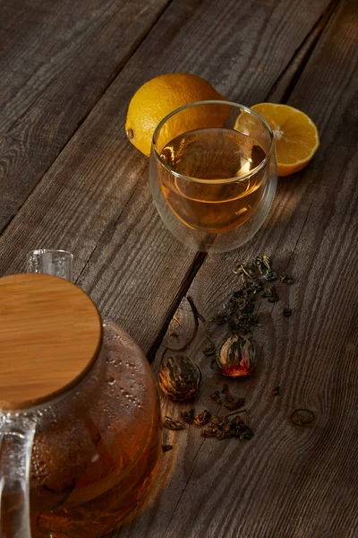 Limones, bolas de té y tetera transparente con vaso de té chino en flor sobre mesa de madera - foto de stock