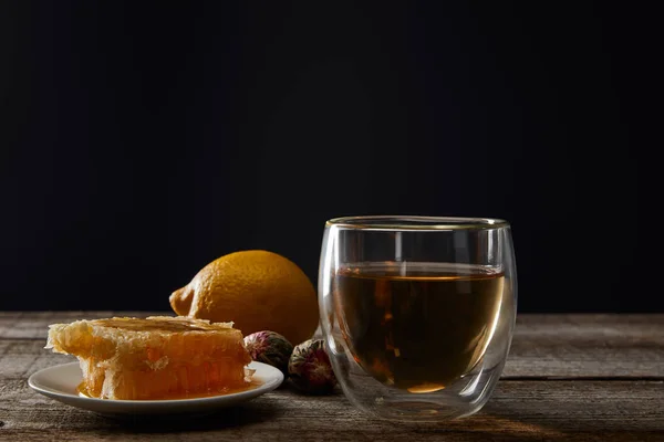 Transparent glass with blooming tea, honeycomb and lemon on wooden table isolated on black — Stock Photo