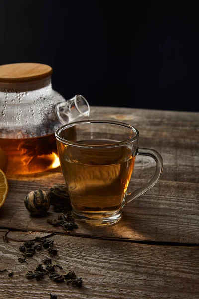 Tetera transparente y taza con té tradicional en flor sobre mesa de madera aislada en negro - foto de stock