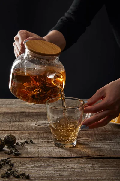 Abgeschnittene Ansicht der jungen Frau gießt blühenden Tee aus Teekanne in Tasse isoliert auf schwarz — Stockfoto