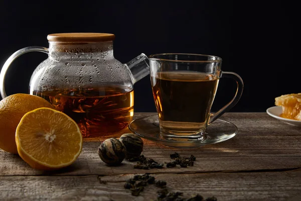 Teiera trasparente e tazza con tè fiorito tradizionale su tavolo di legno con limoni isolati su nero — Foto stock