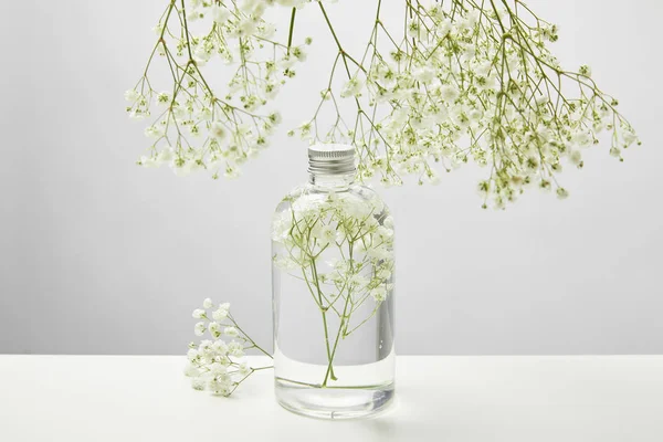 Natural beauty product in transparent bottle and white wildflowers around on grey background — Stock Photo