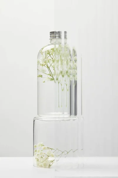 Produit cosmétique bio en flacon transparent avec fleurs sauvages derrière verre sur table blanche sur fond gris — Photo de stock