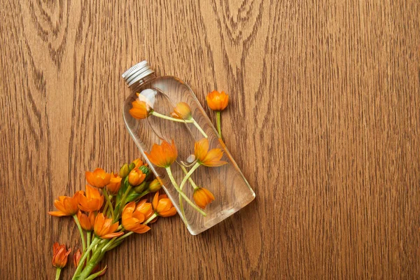 Вид на прозрачную бутылку с органической косметикой рядом с оранжевыми цветами на деревянном фоне — стоковое фото