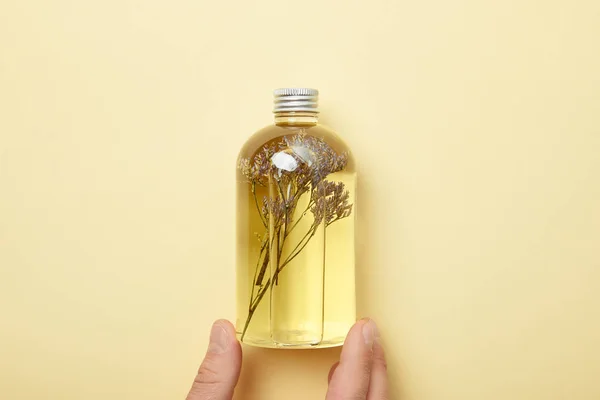 Vista recortada del hombre sosteniendo botella con producto de belleza natural y hierbas sobre fondo amarillo - foto de stock