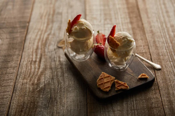 Вкусное мороженое в стаканах с клубникой и кусочками вафель на раскройке доски на деревянном столе — стоковое фото