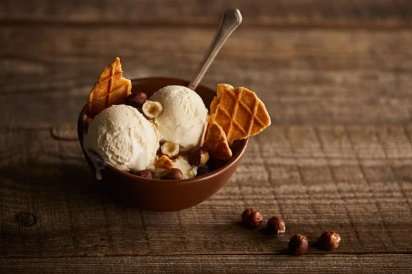 Delicious ice cream with pieces of waffle, spoon and hazelnuts in bowl on wooden surface — Stock Photo