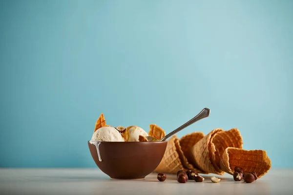 Bowl of delicious ice cream with waffle cones, hazelnuts and spoon on blue with copy space — Stock Photo