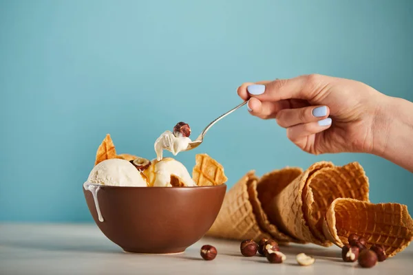 Cropped view of woman with spoon, bowl of ice cream, hazelnuts and waffle cones on blue — Stock Photo