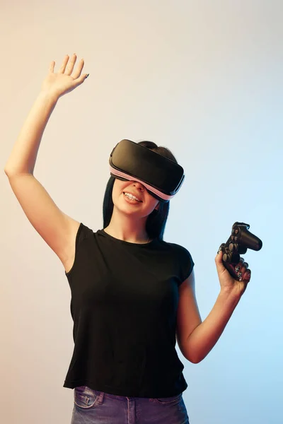 KYIV, UKRAINE - APRIL 5, 2019: Cheerful woman holding joystick while wearing virtual reality headset and waving hand on beige and blue — Stock Photo