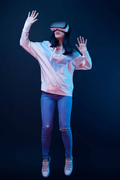 Morena mujer joven haciendo gestos mientras usa auriculares de realidad virtual en azul - foto de stock