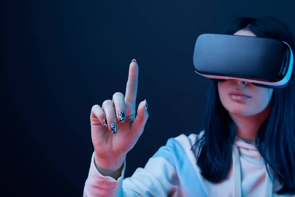 Enfoque selectivo de la joven morena con auriculares de realidad virtual y apuntando con el dedo en azul - foto de stock