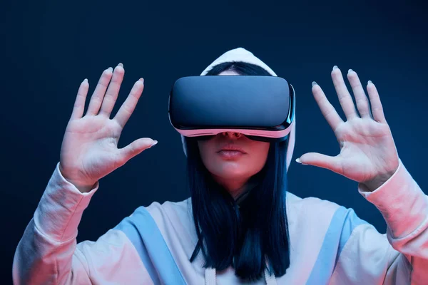 Selective focus of brunette girl in hood gesturing while virtual reality headset on blue — Stock Photo