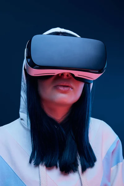 Vista basso angolo di bruna giovane donna nel cappuccio utilizzando cuffia realtà virtuale su blu — Foto stock