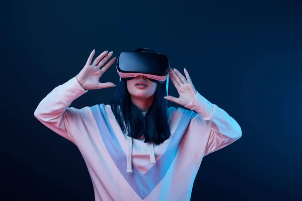 Mujer joven sorprendida haciendo gestos mientras usa auriculares de realidad virtual en azul - foto de stock