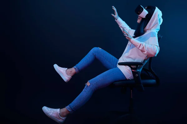 Весела жінка жестикулює і використовує гарнітуру віртуальної реальності, сидячи на стільці на синьому — стокове фото