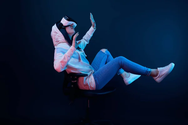 Surpris jeune femme gestuelle et en utilisant casque de réalité virtuelle tout en étant assis sur la chaise sur bleu — Photo de stock