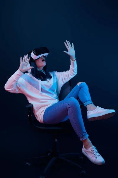 Shocked young woman gesturing and using virtual reality headset while sitting on chair on blue — Stock Photo