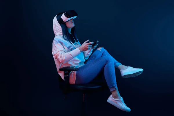 KYIV, UKRAINE - APRIL 5, 2019: Brunette woman sitting on chair and playing video game while wearing virtual reality headset on blue — Stock Photo
