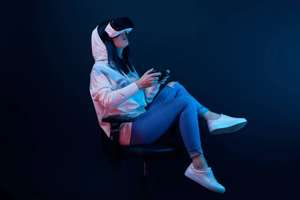 KYIV, UCRAINA - 5 APRILE 2019: Donna bruna seduta sulla sedia e che gioca ai videogiochi indossando cuffie di realtà virtuale su blu — Foto stock