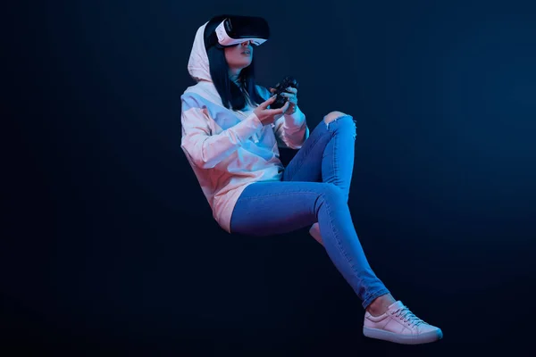 KYIV, UCRANIA - 5 DE ABRIL DE 2019: Mujer joven sorprendida con auriculares de realidad virtual levitando y sosteniendo el joystick en azul - foto de stock