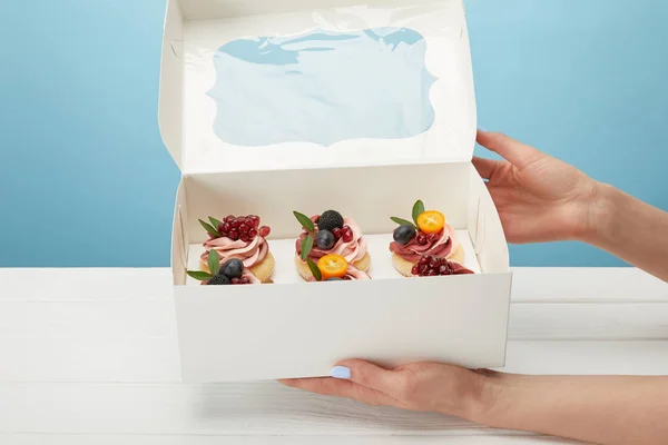 Partial view of woman holding box with cupcakes isolated on blue — Stock Photo