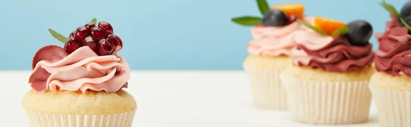 Panoramic shot of cupcakes with cream and garnet on white surface isolated on blue — Stock Photo