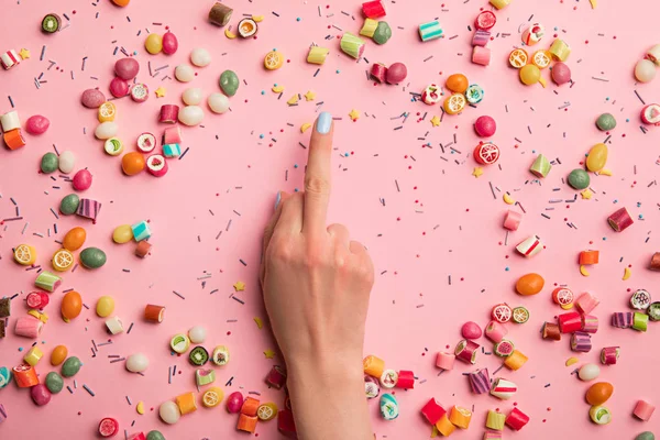 Cropped view of woman showing middle finger near multicolored candies and sprinkles scattered on pink background — Stock Photo