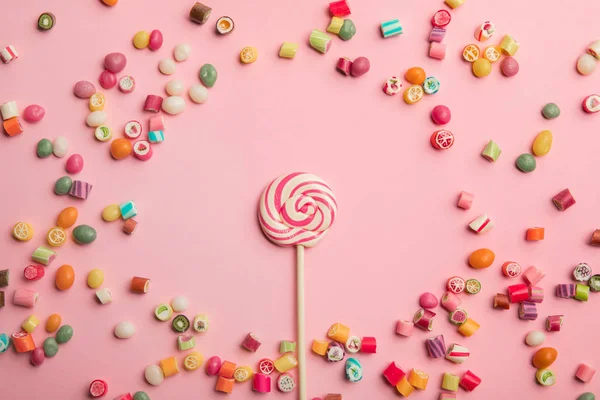 Vue du dessus de délicieuse sucette tourbillonnante sur bâton de bois avec des bonbons au caramel éparpillés autour sur fond rose — Photo de stock