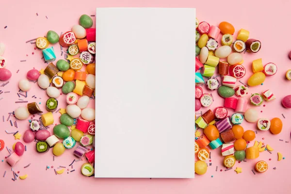 Vista superior de deliciosos caramelos multicolores y tarjeta blanca con espacio de copia sobre fondo rosa - foto de stock