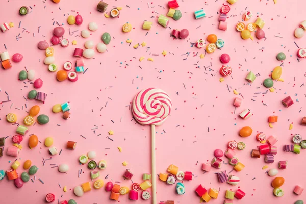 Draufsicht auf köstlichen Wirbel-Lutscher auf Holzstab mit verstreuten Karamellbonbons und Streuseln auf rosa Hintergrund — Stockfoto