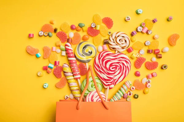 Вид сверху на вкусные разноцветные конфеты, разбросанные из бумажного пакета на ярко-желтом фоне — стоковое фото