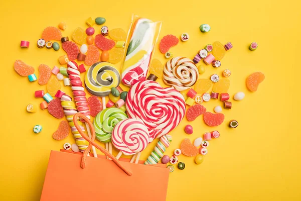 Вид зверху смачні різнокольорові цукерки, розкидані з паперового мішка на яскраво-жовтому тлі — стокове фото