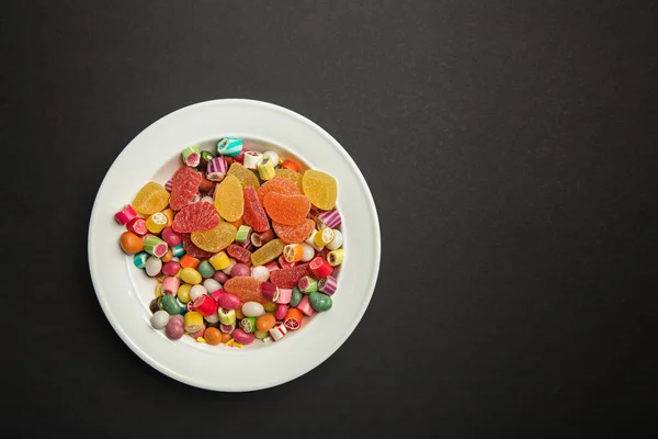 Vista superior de deliciosos doces de caramelo multicoloridos e geleias cítricas em placa branca no fundo preto com espaço de cópia — Fotografia de Stock