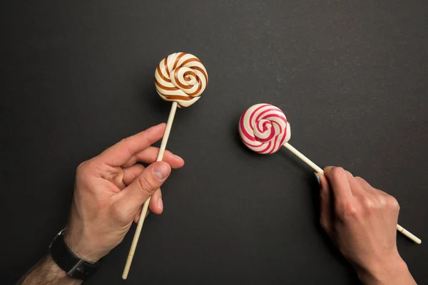 Vista recortada del hombre y la mujer sosteniendo piruletas dulces en palos de madera sobre fondo negro - foto de stock