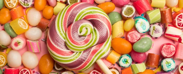 Tiro panorâmico de pirulito redondo multicolorido em pau de madeira perto de doces de caramelo de frutas no fundo rosa — Fotografia de Stock