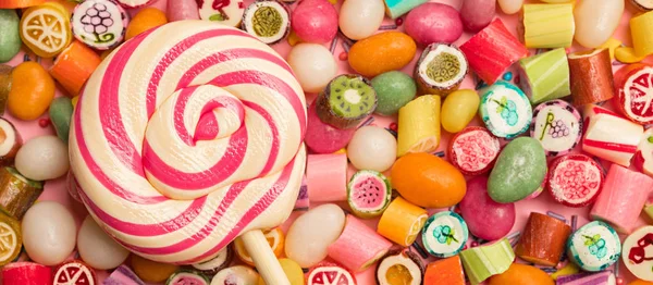 Tiro panorâmico de pirulito redondo brilhante em pau de madeira perto de doces de caramelo de frutas no fundo rosa — Fotografia de Stock